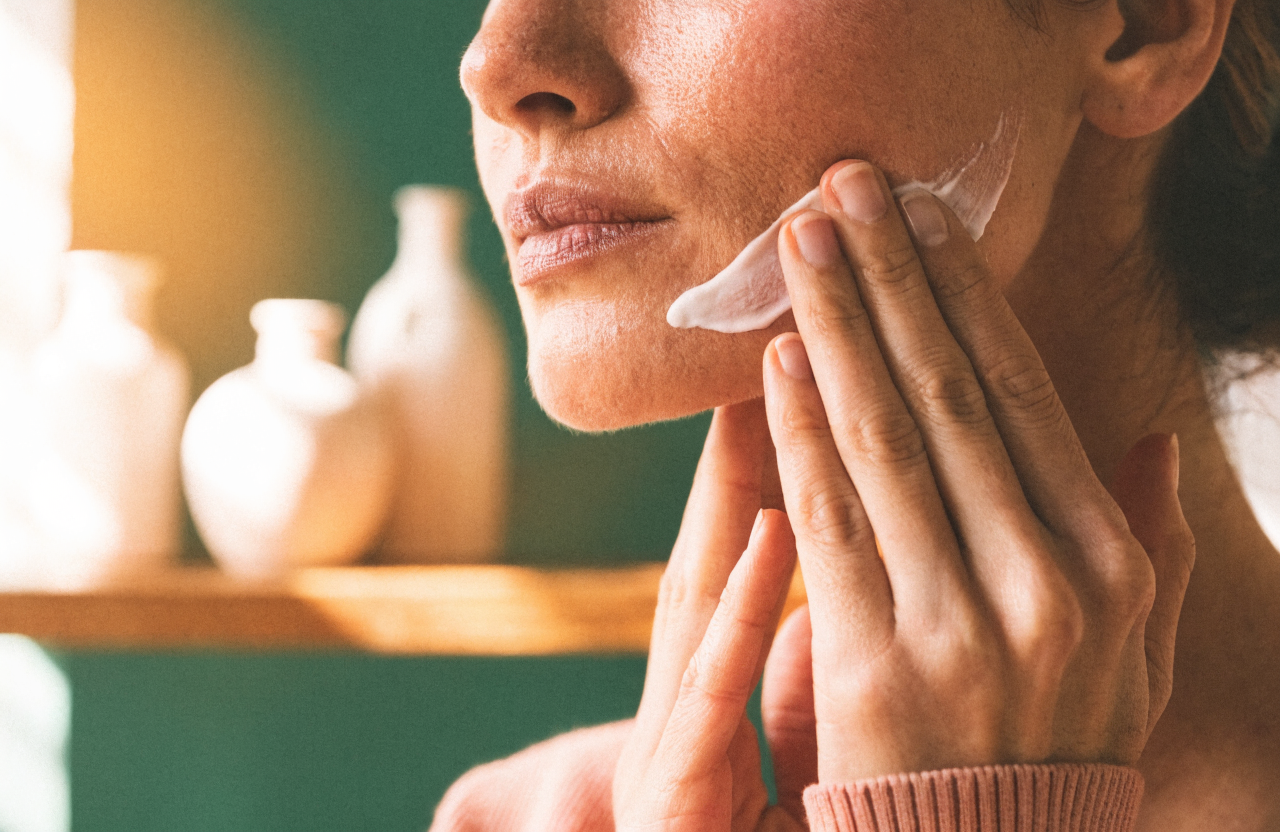 Cream being smoothed along a jawline, with plain bottles on a shelf behind.