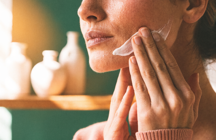 Cream being smoothed along a jawline, with plain bottles on a shelf behind.