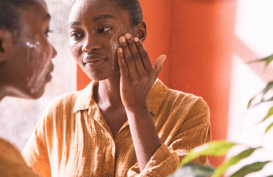 A woman smoothing serum along her cheek at a mirror in warm morning light.
