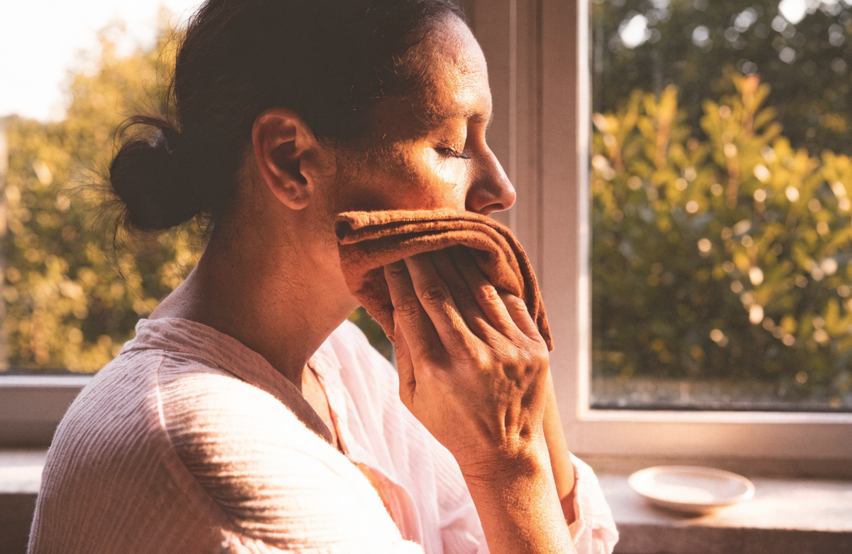 A woman pressing a folded linen cloth to her cheek by a sunlit window.