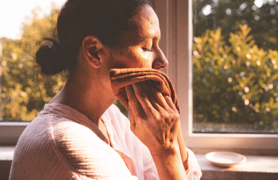 A woman pressing a folded linen cloth to her cheek by a sunlit window.